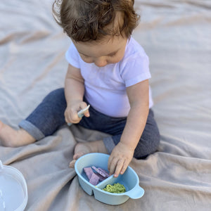 Baby holds baby spoon in right hand, and reaches baby crackers in a blue divided bowl for travel.